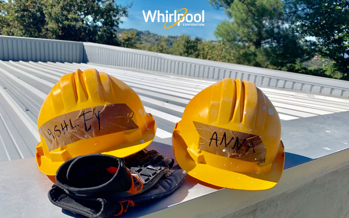 Two yellow hard hats with work gloves, resting on a roof, one has name Ashley, and one has name Anna