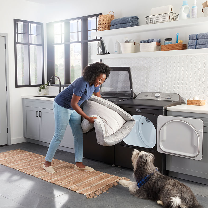 Maytag Pet Pro laundry pair with woman putting a dog bed into the washer with a dog looking on