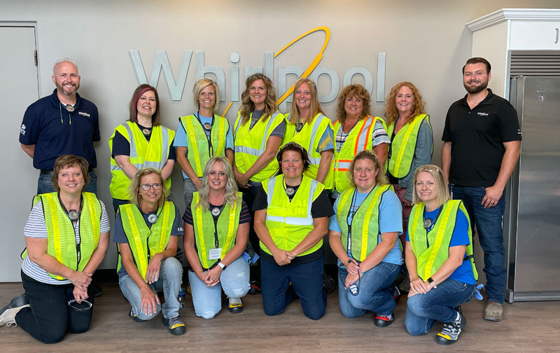 Twelve female educators and two male manufacturing leaders pose for a group photo. All teachers are wearing bright yellow safety vests. All are smiling.